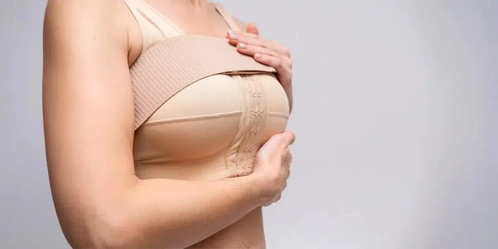 Woman wearing a post-surgical compression bra, gently supporting her chest to aid in breast augmentation recovery.