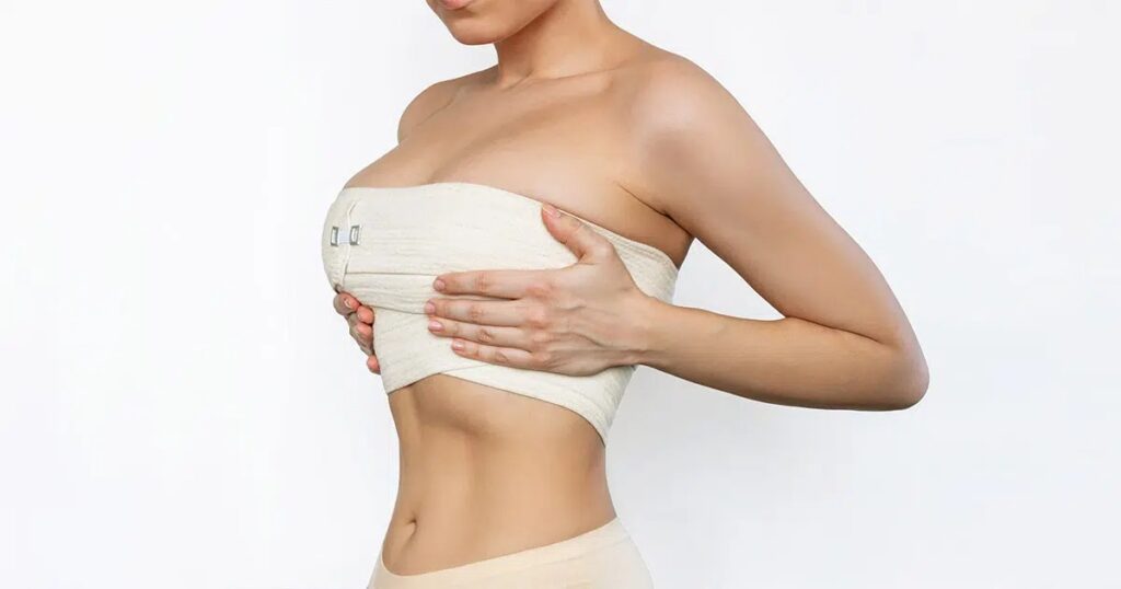 Woman wearing a post-surgical compression garment, gently holding her chest to manage swelling and support healing after breast augmentation surgery.