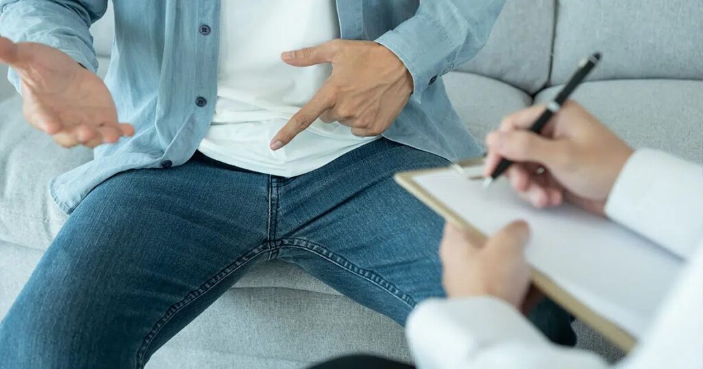 Man sitting on a couch gesturing toward his lower abdomen during a medical consultation, representing discussion of excess fat in the pubic area and options like male pubic liposuction.