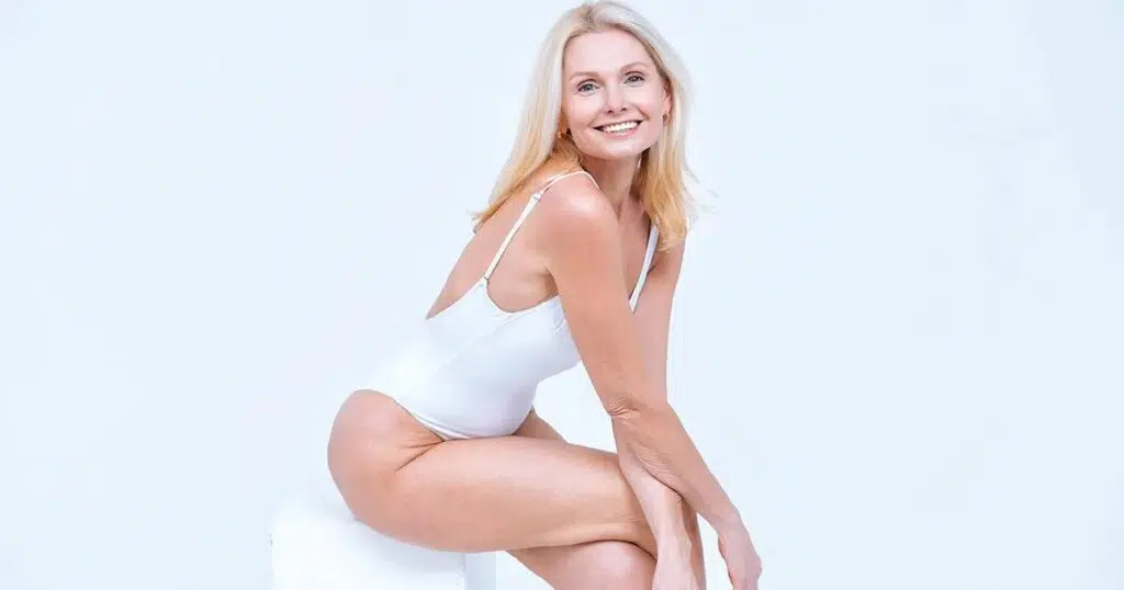 Confident woman smiling in a white bodysuit while seated, representing restored body confidence and healing after mommy makeover recovery.