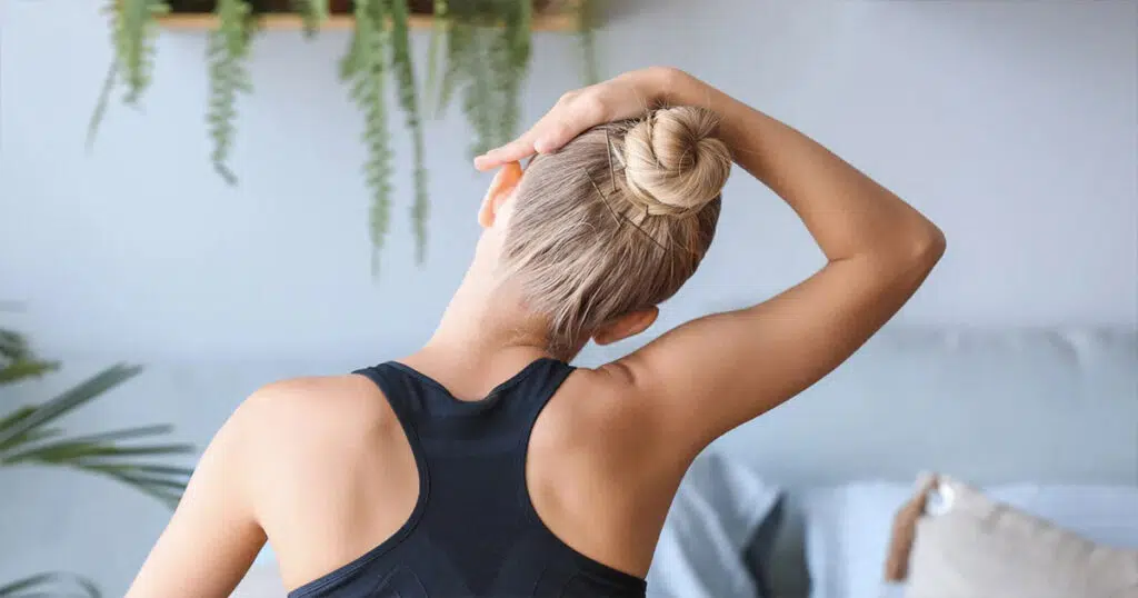 Woman performing a seated neck stretch with one hand over her head, demonstrating a mobility exercise commonly used to reduce tension and improve posture in buffalo hump treatment.