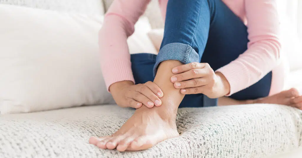 Woman sitting on a couch gently holding her ankle, illustrating concern or discomfort related to cankles or lower leg swelling.