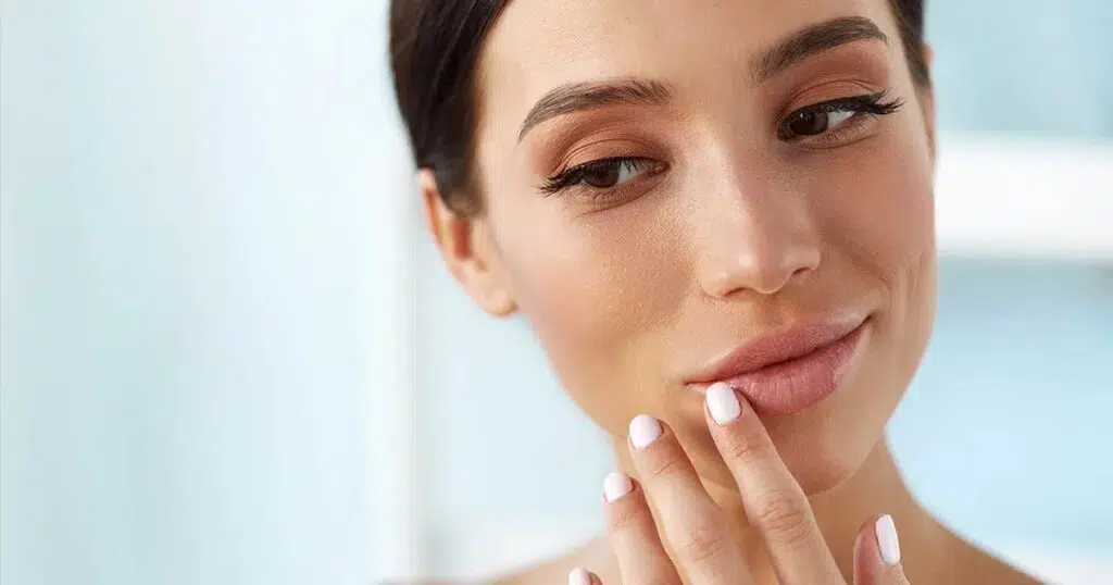 Woman gently touching her lips while looking in the mirror, representing the natural fullness and contour enhancement achieved through lip fat transfer.