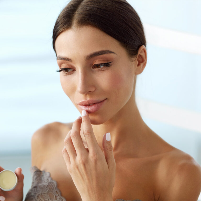 Woman touching her lower lip with one finger while holding a small open pot of balm