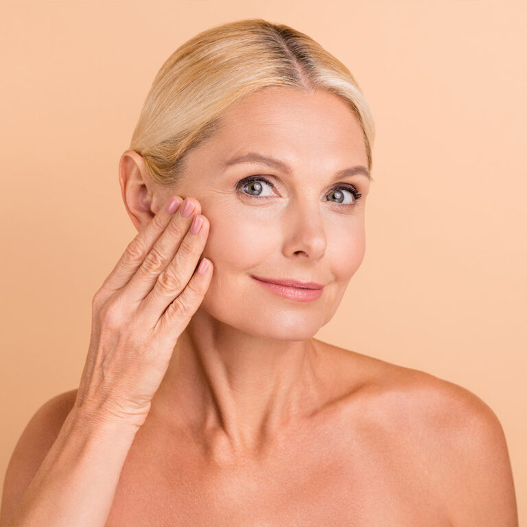 Older woman with bare shoulders resting a flat hand against her cheek and jaw, beige background