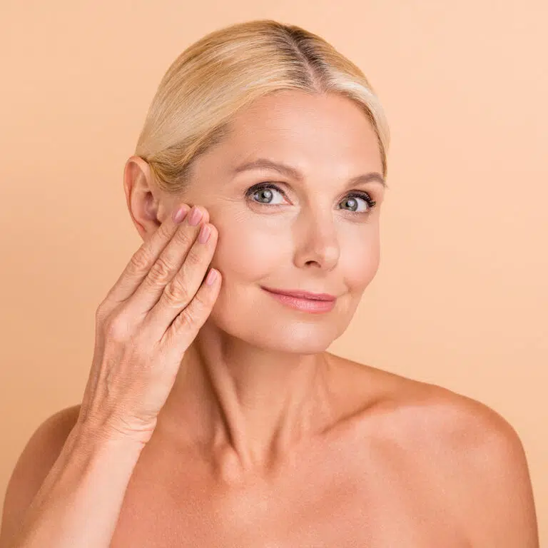 Older woman with bare shoulders resting a flat hand against her cheek and jaw, beige background