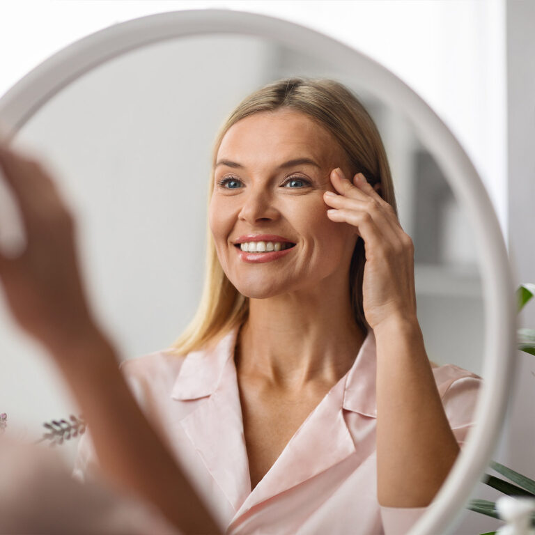 Woman in a pink satin shirt seen in a round mirror, fingertips at the outer corner of her eye