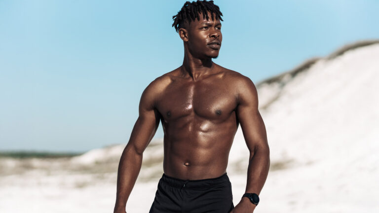 Fit shirtless man with a defined abdomen standing outdoors against white sand dunes